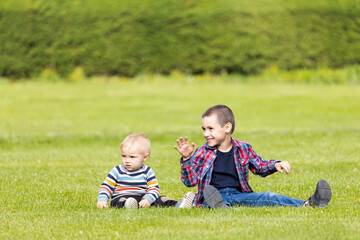 Fototapeta premium Two cheerful boy brothers of different ages play fun, sit on the grass on a warm summer day. The concept of friendship of brothers and happy childhood.