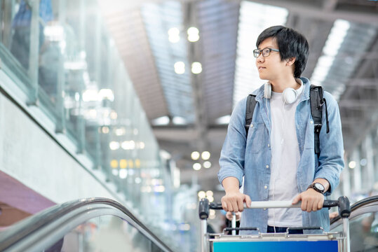 Vacation trip or travel abroad concept. Asian man tourist in casual clothing carrying backpack walking with trolley cart on travelator in international airport terminal.