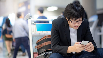 Business travel concept. Smart Asian businessman in formal suit using smartphone for airline online check-in while sitting with suitcase luggage on trolley cart at departure hall in airport terminal.