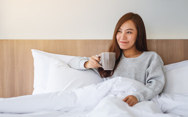Fototapeta na wymiar A beautiful asian woman drinking hot coffee while lying on a white cozy bed at home in the morning