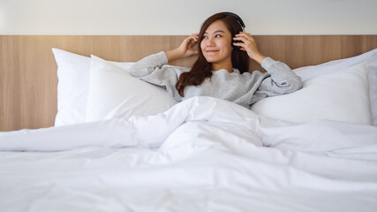 A beautiful asian woman enjoy listening to music with headphone while lying on a white cozy bed at home