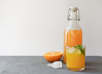 orange juice in a bottle and in a glass with ice cubes. orange and mint as summer cocktail ingredients. fresh cold drink for summer hot. copy space and grey background. 