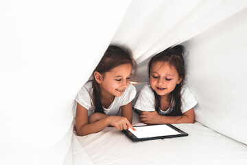 Two beautiful little sisters lying in the bed and look at the screen of a tablet, smart technology