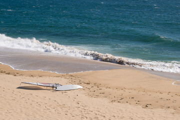 Windsurfing board lying on sandy beach on sunny day in summer with ocean waves breaking behind