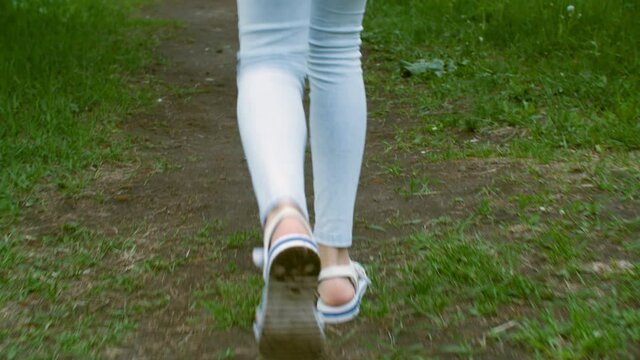 Teenager girl legs in blue leggings and sandals walking then running on grass lawn. Walking female legs in summer sandals on green grass back view