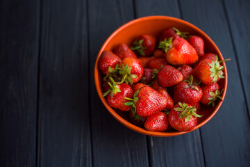 Close-up fresh ripe delicious strawberries in a orange bowl plate on wooden black table background. Antioxidant nutrition, healthy organic food concept. Copyspace. Flat lay. Daylight