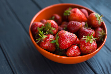 Close-up fresh ripe delicious strawberries in a orange bowl plate on wooden black table background. Antioxidant nutrition, healthy organic food concept. Copyspace. Flat lay. Daylight