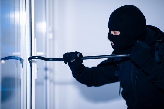 Robber in black balaclava cracking door with crowbar