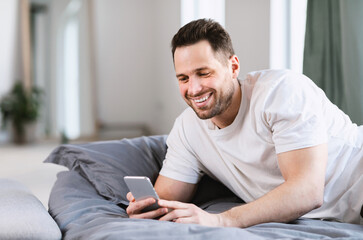 Fototapeta premium Happy Man Using Smartphone Lying In Bed At Home