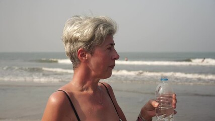 Mature woman senior citizen drinks clear water from a bottle on nature.