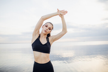 young woman in black sportswear stretching hands by the river. Fitness girl with hands raised above her head stretching outdoors on sunny summer day