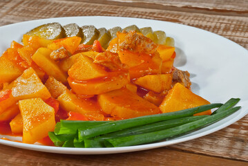 Potatoes in a white plate on a wooden table. Sliced potatoes with meat and onions. Boiled potatoes with orange sauce. Food in utensils on the background of an old pink board.