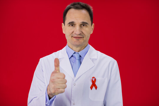 Handsome serious male senior age doctor in white lab coat with red ribbon on coat shows like sign with thumb up on red background, copy space. Symbol of HiV Aids struggle, awareness, world aids day - Powered by Adobe