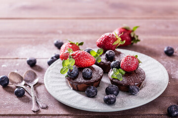 Chocolate cakes (cupcakes) are on the table. Cupcakes decorated  blueberries, strawberries and green leaves mint on top. Beautiful and elegant serving with white dishes
