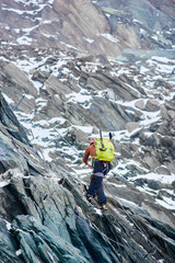 Back view of male mountaineer with backpack using fixed rope to climb high rocky mountain. Climber ascending alpine ridge and trying to reach mountaintop. Concept of alpinism and alpine rock climbing.