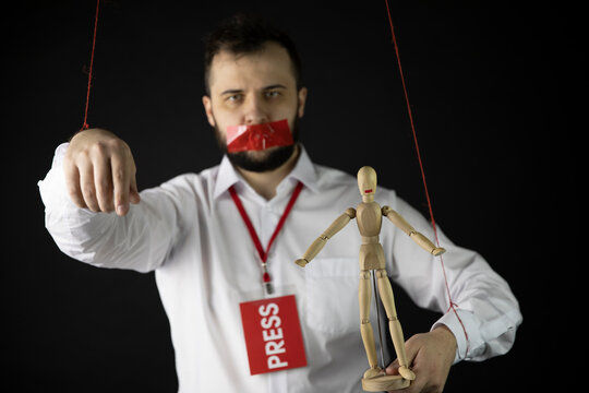 Journalist With Badge Press Holds Hands Tied With Ropes Like Marionette With Wrapping Mouth By Adhesive Tape And Dummy In Hand Isolated On Dark Background. Media Freedom Concept