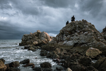 man sitting on top of the rock at sea shore with storm clouds in background during windy day in the beginning of rainy season 