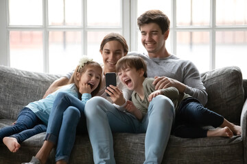 Overjoyed young family with small children sit relax on sofa at home laugh watching funny video on...