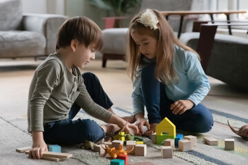 Fototapeta premium Concentrated little brother and sister sit on warm floor in living room play with building bricks or blocks, small preschooler boy and girl children have fun engaged in game with wooden toys at home