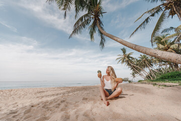 Fototapeta na wymiar Beautiful woman with long blond hair is holding pineapple fruit on the beach, sitting on the background of ocean and coconut palm trees. Vacation on tropical island