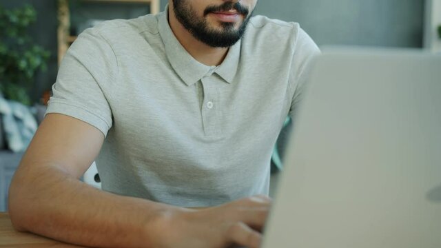 Tilt-up portrait of good-looking bearded Arab man working with laptop computer in apartment typing sitting at wooden table in cozy room. People and freelance work concept.