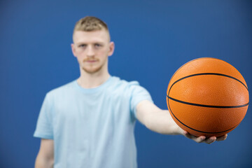 Young caucasian bearded student in blue T-shirt holds basketball on outstretched arm on isolated dark blue background. Sport, youth, activity, health, sport section concept. Focus on ball