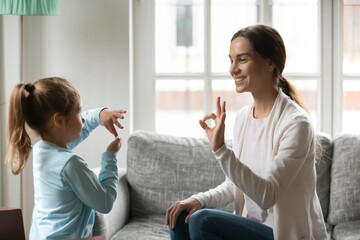 Smiling young Caucasian mom and little daughter make hand gesture learn speak sign language at...
