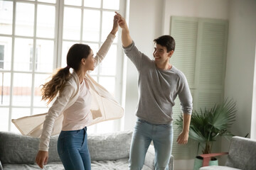 Happy young Caucasian couple dance swirl sway together in living room, smiling millennial man and...