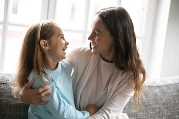 Happy young Caucasian mom and little preschooler daughter sit on couch at home hugging cuddling, smiling mother or nanny rest on sofa embrace have fun play with small girl child, show love and care