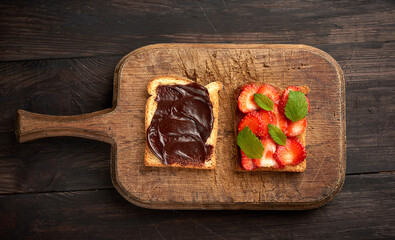 two toasts with chocolate paste and fresh ripe red strawberries on a brown wooden board
