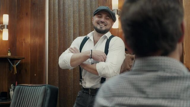 Brutal Barber in a fashionable cap and with a beard conducts a dialogue with a client in a barbershop. Stylishly dressed Barber talks to the client and laughs.