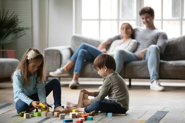 Little preschooler children sit on warm floor in living room play construct with colorful building blocks bricks together, young parents rest on sofa in background, Caucasian family relax at home