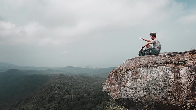 Backpacker on the rock. Tired hiker with backpack sits on the top of mountain. He has no water and trying to drink drop of water from empty bottle.