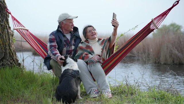 cheerful elderly husband and wife have fun sitting in a hammock by river with a dog, hell like a prod and spend time together talking on a cell phone using a video call in while relaxing