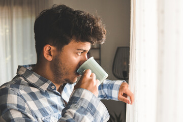 Attractive young man takes a sip of coffee. He's looking out the window.
