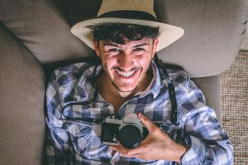 Attractive young man laughs happily as he watches the camera lying on the couch. He holds a camera...