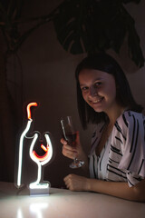 Beautiful girl with a glass of wine on the background of a neon sign of a wine bottle and a glass. Trendy stylish. Glowing life. Modern style. Neon sign.