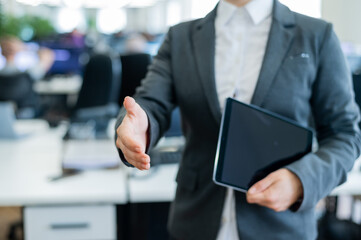 Business woman in a jacket and white shirt holding out a hand for a greeting. A faceless business woman holds a digital tablet and shakes her hand as an agreement on a successful transaction.