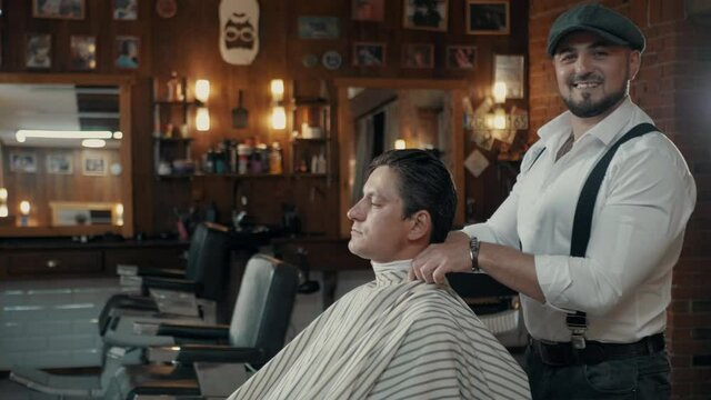 A stylishly dressed barber prepares the client for a haircut. Brutal Barber with a beard and a cap is dancing.