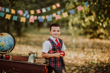 Young boy in school uniform posing with newton apple near an old wooden desk in the park. Back to...