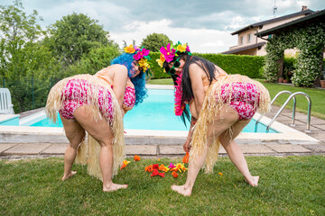 two female friends in Hawaiian costumes are picking flowers from a lawn © Marino Bocelli