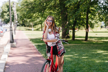 Adorable smiling girl posing in park with bicycle. Outdoor photo of relaxed lady posing on nature background.