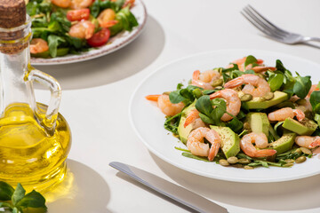 selective focus of fresh green salad with shrimps and avocado on plate near olive oil and cutlery on white background