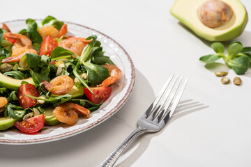 selective focus of fresh green salad with shrimps and avocado on plate near fork and ingredients on white background