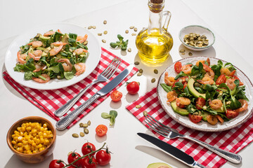 fresh green salad with shrimps and avocado on plates near cutlery on plaid napkins and ingredients on white table