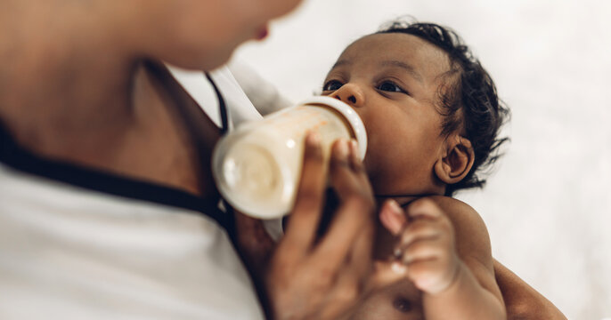 Portrait Of Enjoy Happy Love Family African American Mother Playing With Adorable Little African American Baby.Mom Feeding Bottle Of Milk To Baby Cute Son In A White  Bedroom.Love Of Black Family 