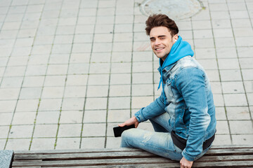 Side view of positive man in earphones smiling at camera while holding smartphone on bench