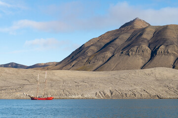 Red sailing ship in front of mountains without vegetation. Summer without snow in the Arctic