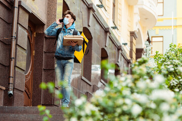 Selective focus of courier in medical mask talking on smartphone and holding pizza boxes on urban street