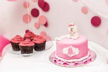 
A pink cake with a white dog figurine next to a cupcake plate for celebrating a dog’s holiday.